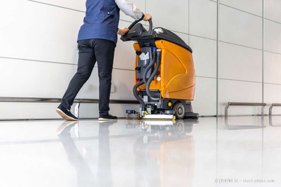 Eine Person reinigt in einem weiß getünchten Raum einen glänzend weißen Boden mit einem orangefarbenen Industriebodenreiniger.