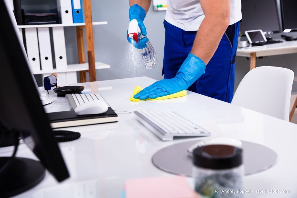 Eine Person mit blauen Handschuhen besprüht und wischt einen Schreibtisch in einem Büro ab.