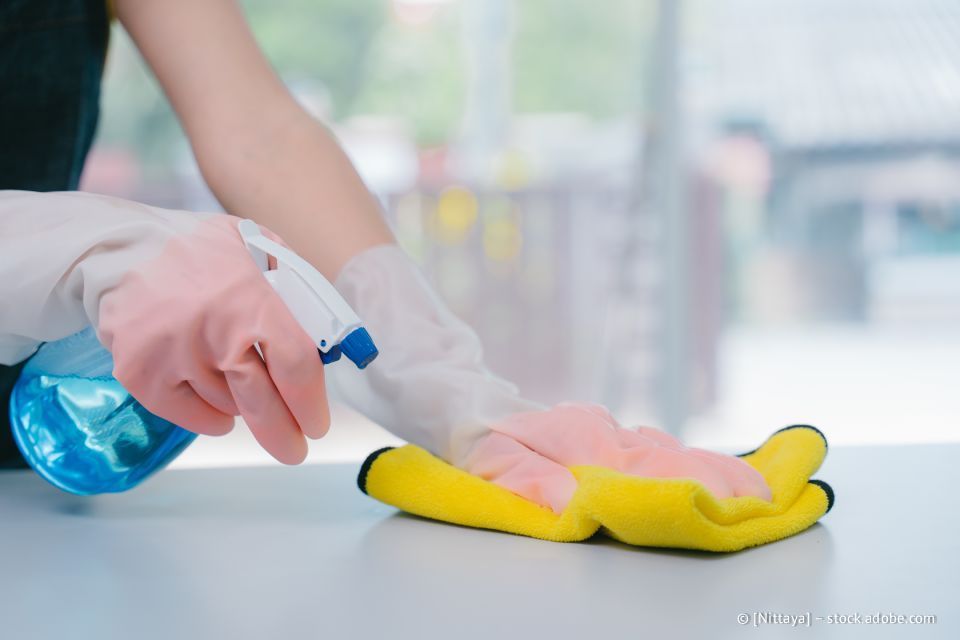 Eine Person mit rosa Handschuhen besprüht und wischt eine weiße Oberfläche mit einem gelben Tuch ab.