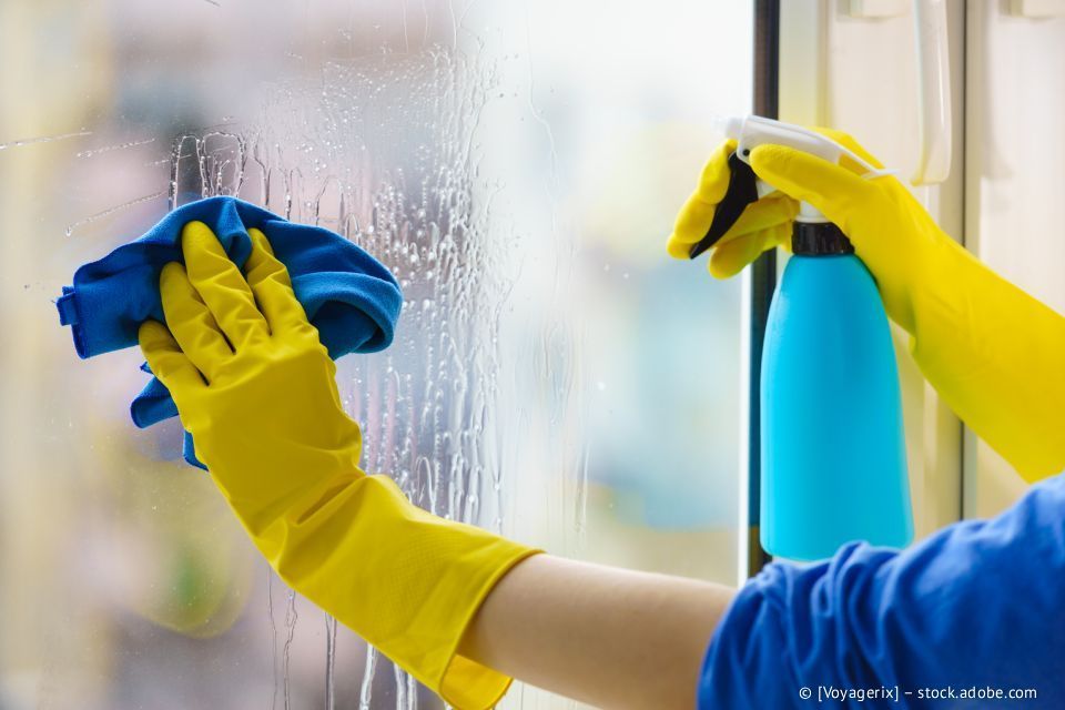 Eine Person mit gelben Handschuhen reinigt ein Fenster mit einer blauen Sprühflasche und einem Tuch.