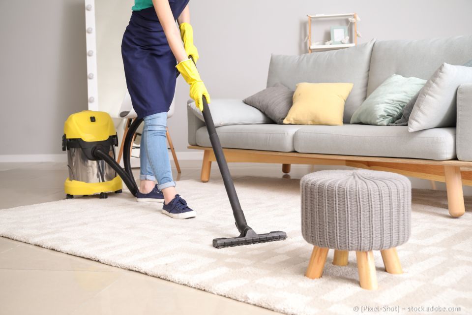 Eine Person saugt mit Handschuhen und Schürze einen hellen Teppich im Wohnzimmer neben einem Sofa und einem Hocker.