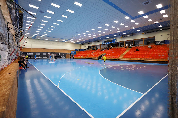 Indoor-Sportarena mit blauem Boden und orangefarbenen Sitzreihen. Zuschauer sind auf dem Spielfeld und auf den Rängen zu sehen.