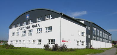 Weißes und graues Sporthallengebäude mit Bogendach; davor grüner Rasen.