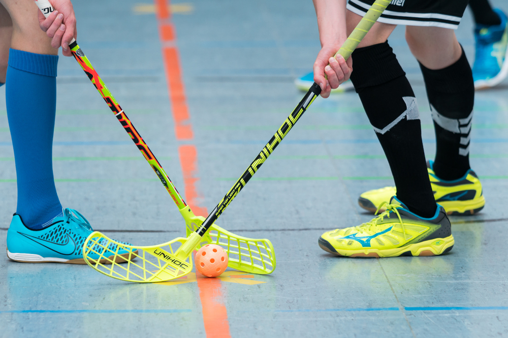 Zwei Spieler stehen sich in einem Floorballspiel gegenüber, ihre Schläger kreuzen sich über einem orangefarbenen Ball.