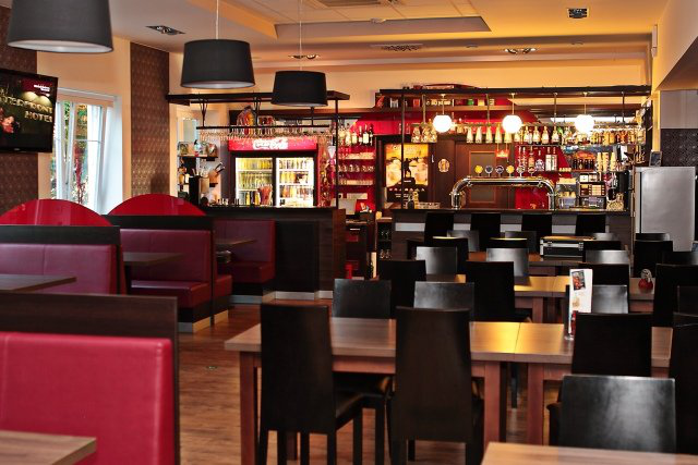 Innenansicht eines Restaurants mit Sitznischen und Tischen. Im Hintergrund ist eine Bar mit Getränken zu sehen. Rot-schwarze Dekoration.