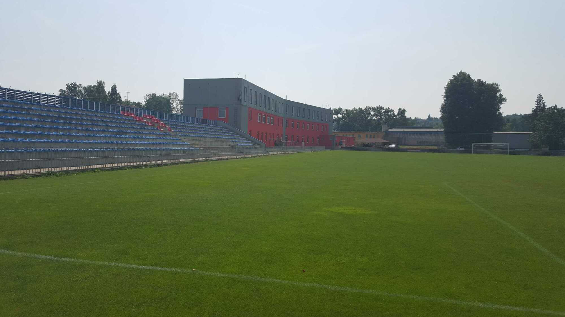 Grüner Fußballplatz mit blauen Tribünen und einem grauen Gebäude.