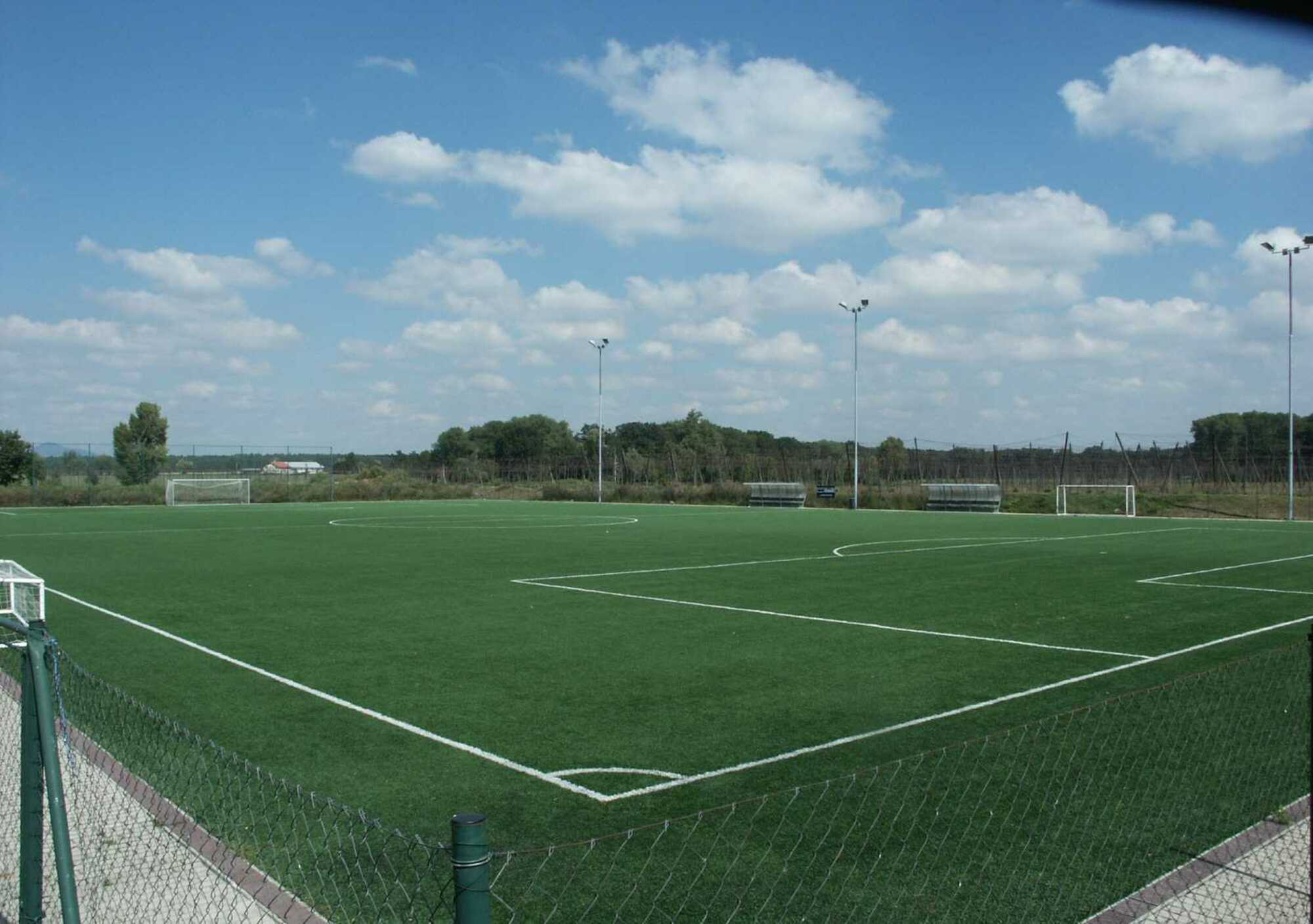 Ein Fußballfeld mit grünem Rasen, weißen Linien, Toren und einem Maschendrahtzaun unter blauem Himmel.