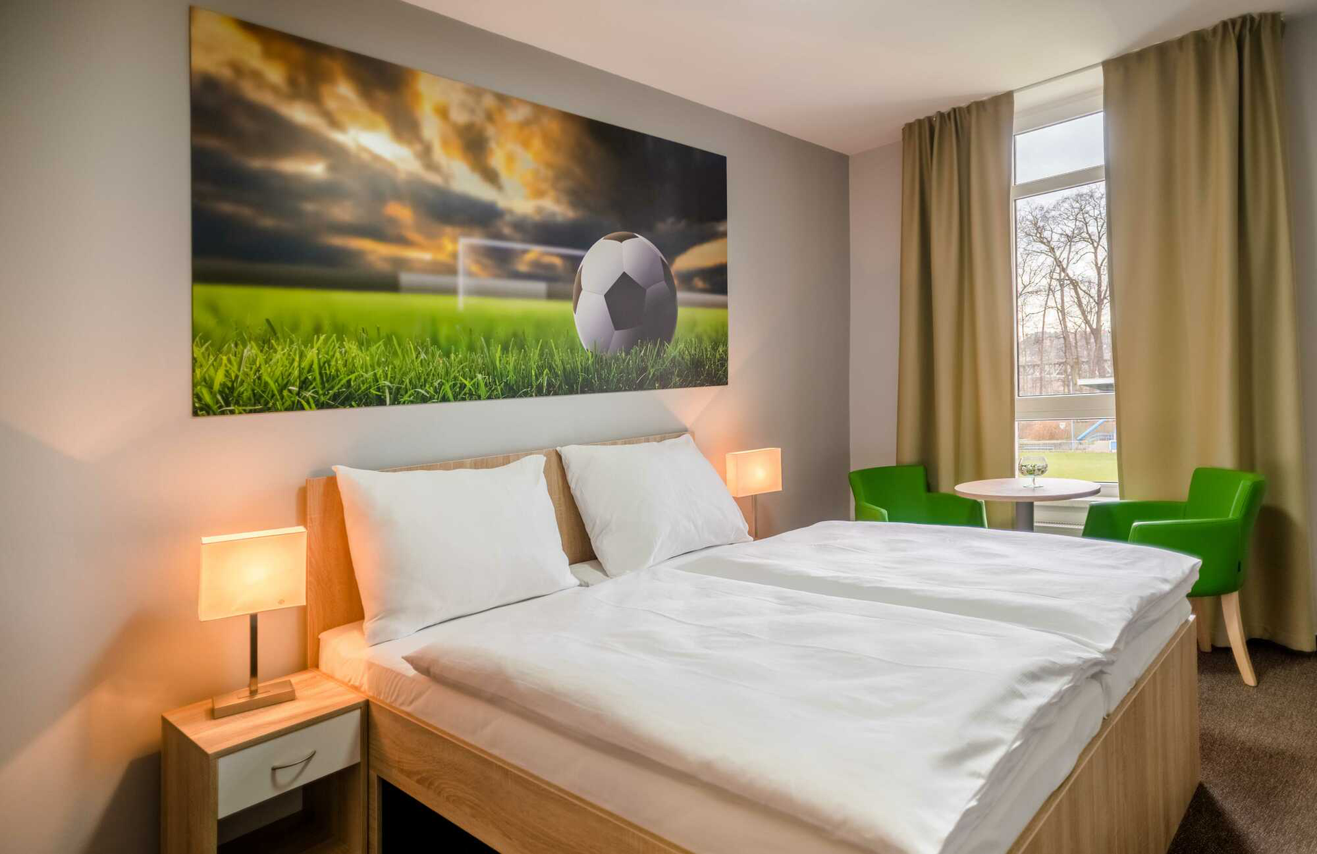 Hotelzimmer mit Doppelbett, Bildern mit Fußballmotiven, Nachttischlampen, Fenster und grünen Stühlen.