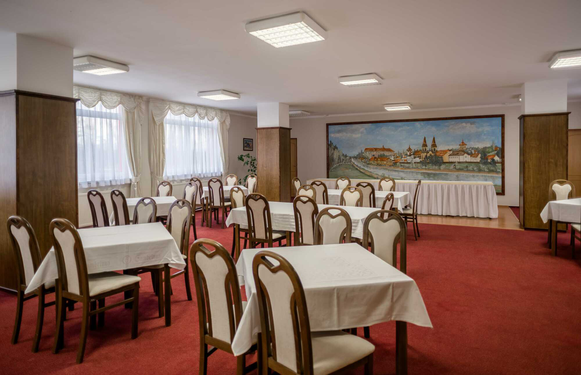 Esszimmer mit weißen Tischdecken, Stühlen, rotem Teppich und einem Wandgemälde.