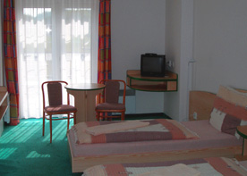 Hotelzimmer mit zwei Betten, kleinem Tisch und Stühlen sowie einem kleinen Fernseher an der Wand. Grüner Teppich, farbenfrohe Vorhänge.