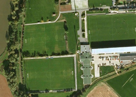 Luftaufnahme eines Sportkomplexes mit mehreren grünen Fußballfeldern, Gebäuden und Parkplätzen.