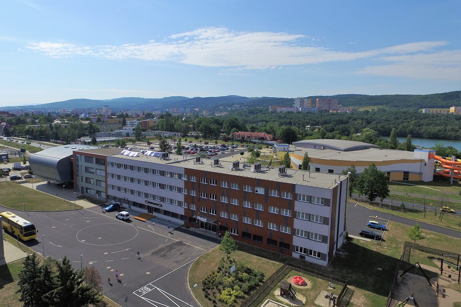 Luftaufnahme eines mehrstöckigen Gebäudes mit rotbrauner Fassade in der Nähe eines Flusses und Bergen unter blauem Himmel.