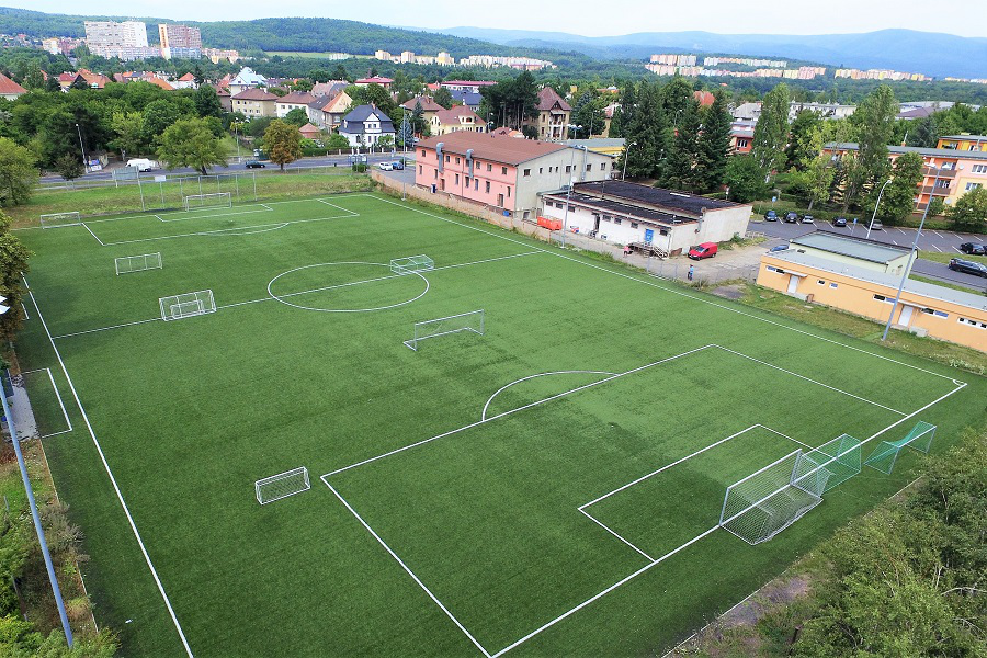 Luftaufnahme eines grünen Fußballfelds mit weißen Markierungen, Toren und Gebäuden im Hintergrund.