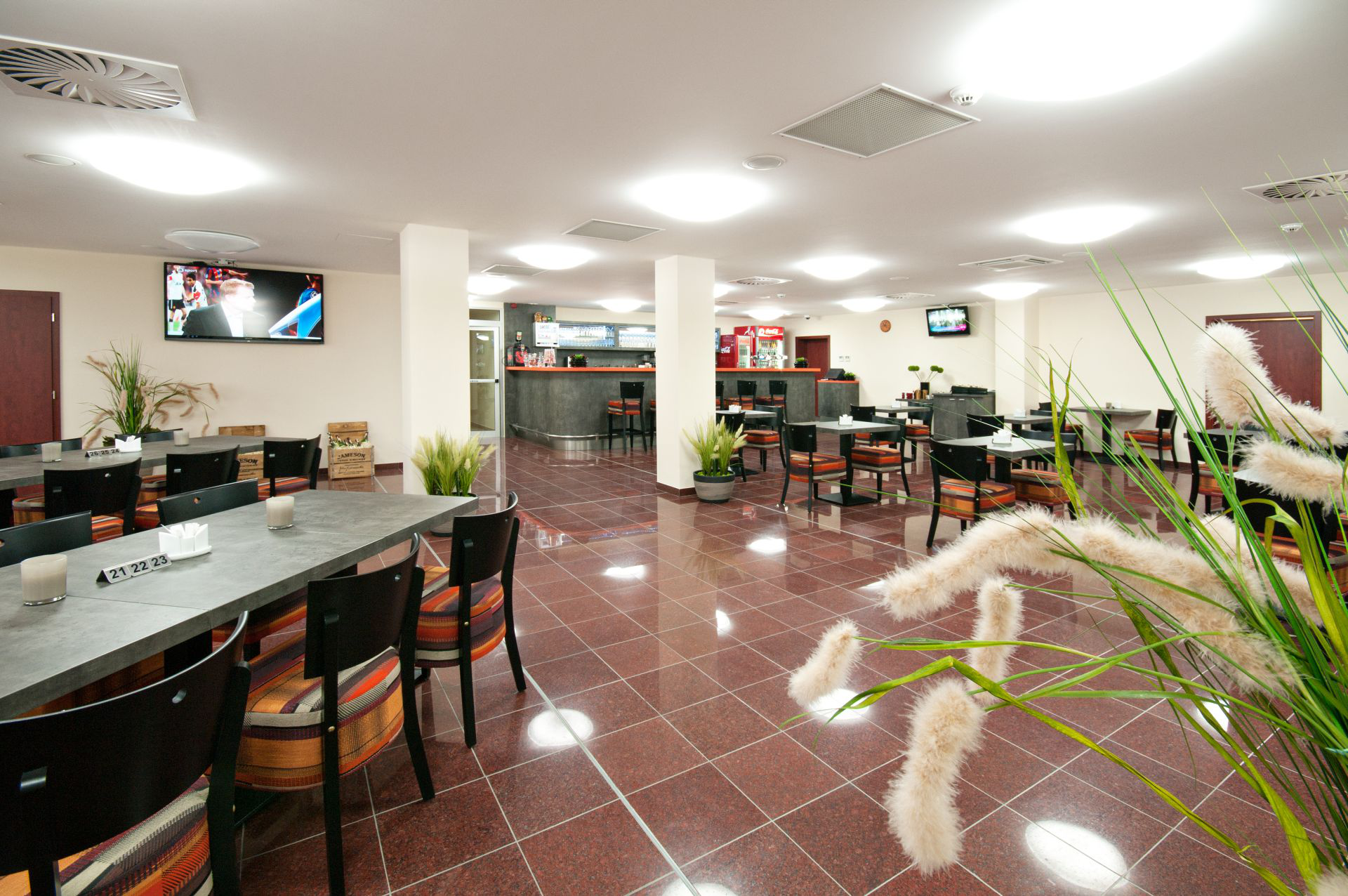 Restaurantinterieur mit Tischen, Stühlen, Bar und Fernsehern. Brauner Fliesenboden, weiße Decke und einige Pflanzen.