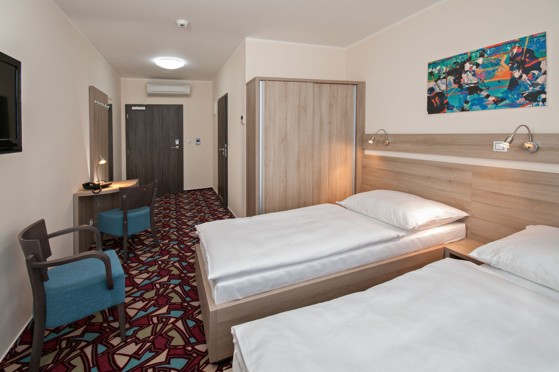 Hotelzimmer mit zwei Einzelbetten, Schreibtisch, Kleiderschrank und Kunstwerken. Rot-braun-blau gemusterter Teppich.