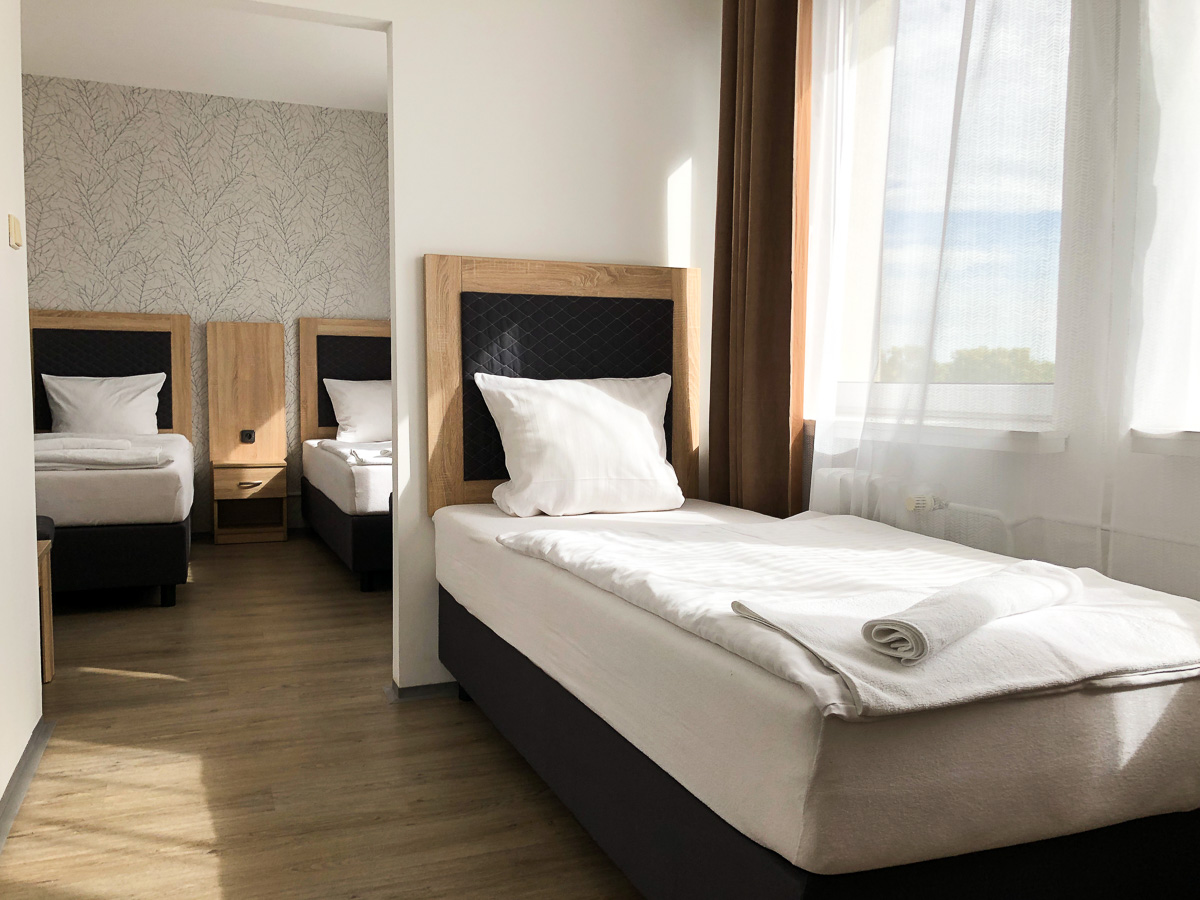 Hotelzimmer mit drei Einzelbetten, zwei im Hintergrund sichtbar, eines im Vordergrund, in der Nähe eines Fensters.
