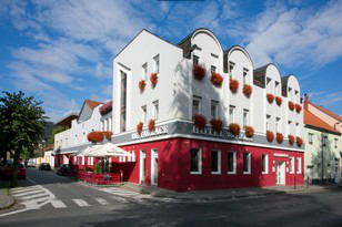 Hotelgebäude, weiße Fassade mit rotem Zierrand, Blumenkästen, Sitzbereich im Freien, blauer Himmel.
