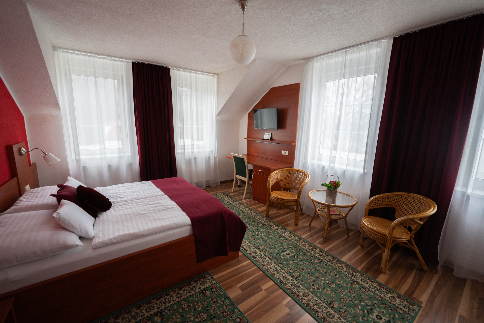 Hotelzimmer mit Bett, Schreibtisch, Korbstühlen und Fenstern mit bordeauxroten Vorhängen.