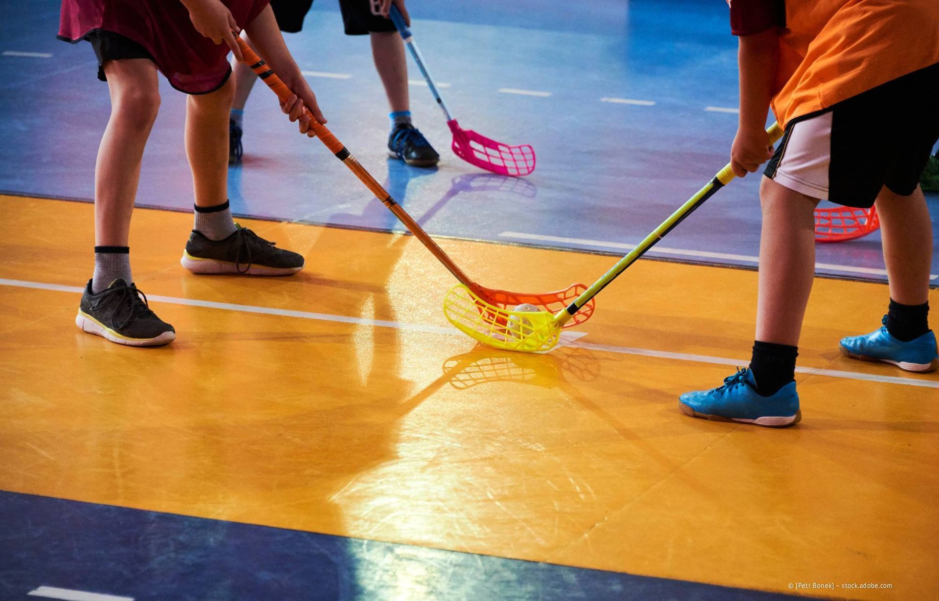 Floorballspieler in Aktion: Einer in weißen Shorts dribbelt den Ball, andere verteidigen. Hallenhockeyfeld.