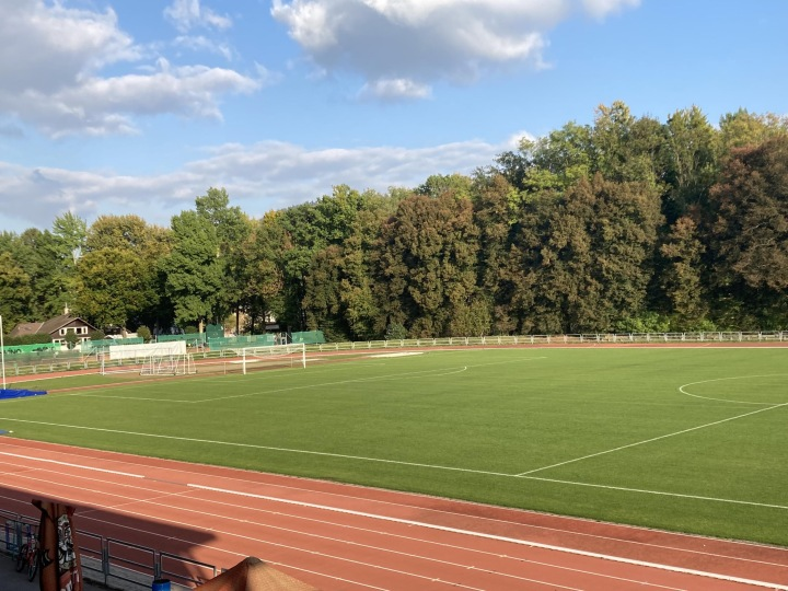 Grüner Fußballplatz mit Laufbahn, umgeben von Bäumen unter blauem Himmel.