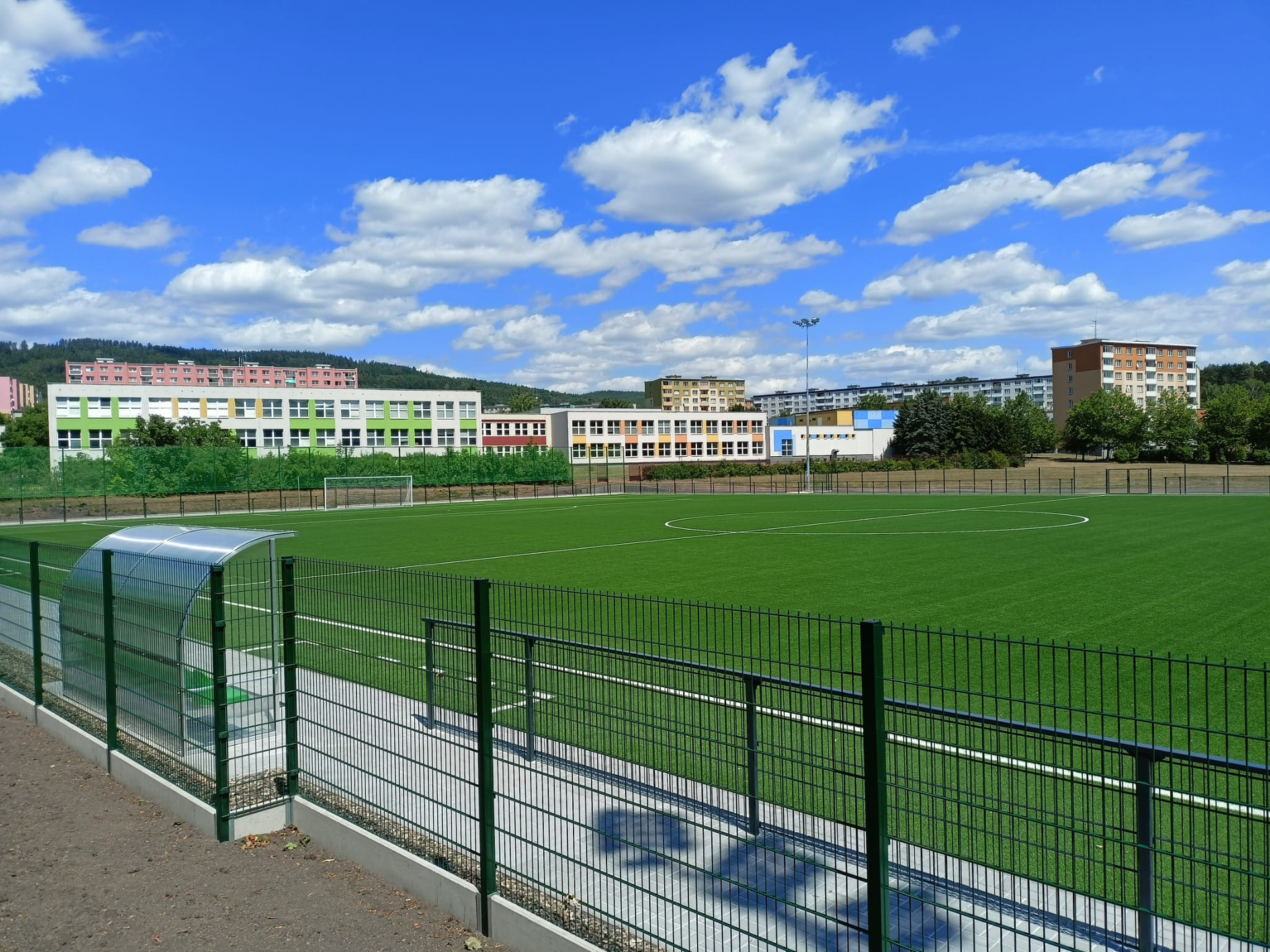 Grüner Kunstrasen-Sportplatz mit Gebäuden und bewölktem, blauem Himmel.