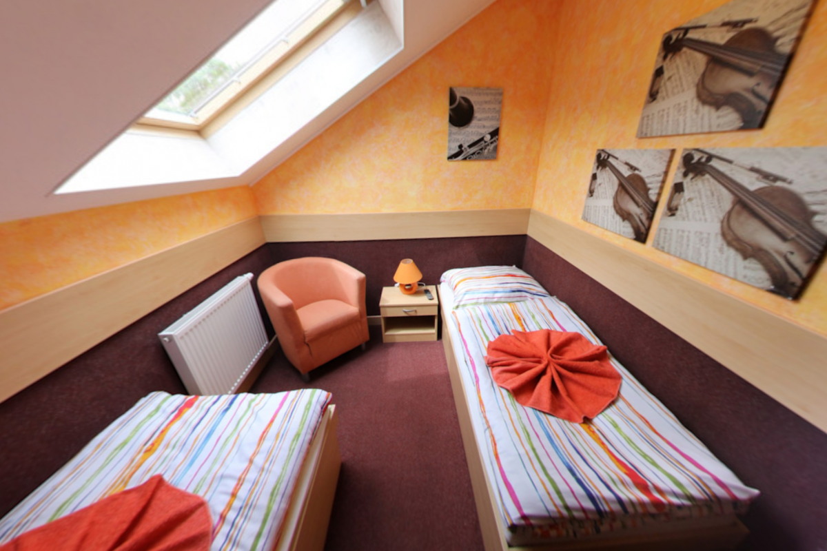Ein kleines Hotelzimmer mit zwei Betten, einem Sessel und einem Dachfenster. Orangefarbene Wände, dunkelroter Teppich.