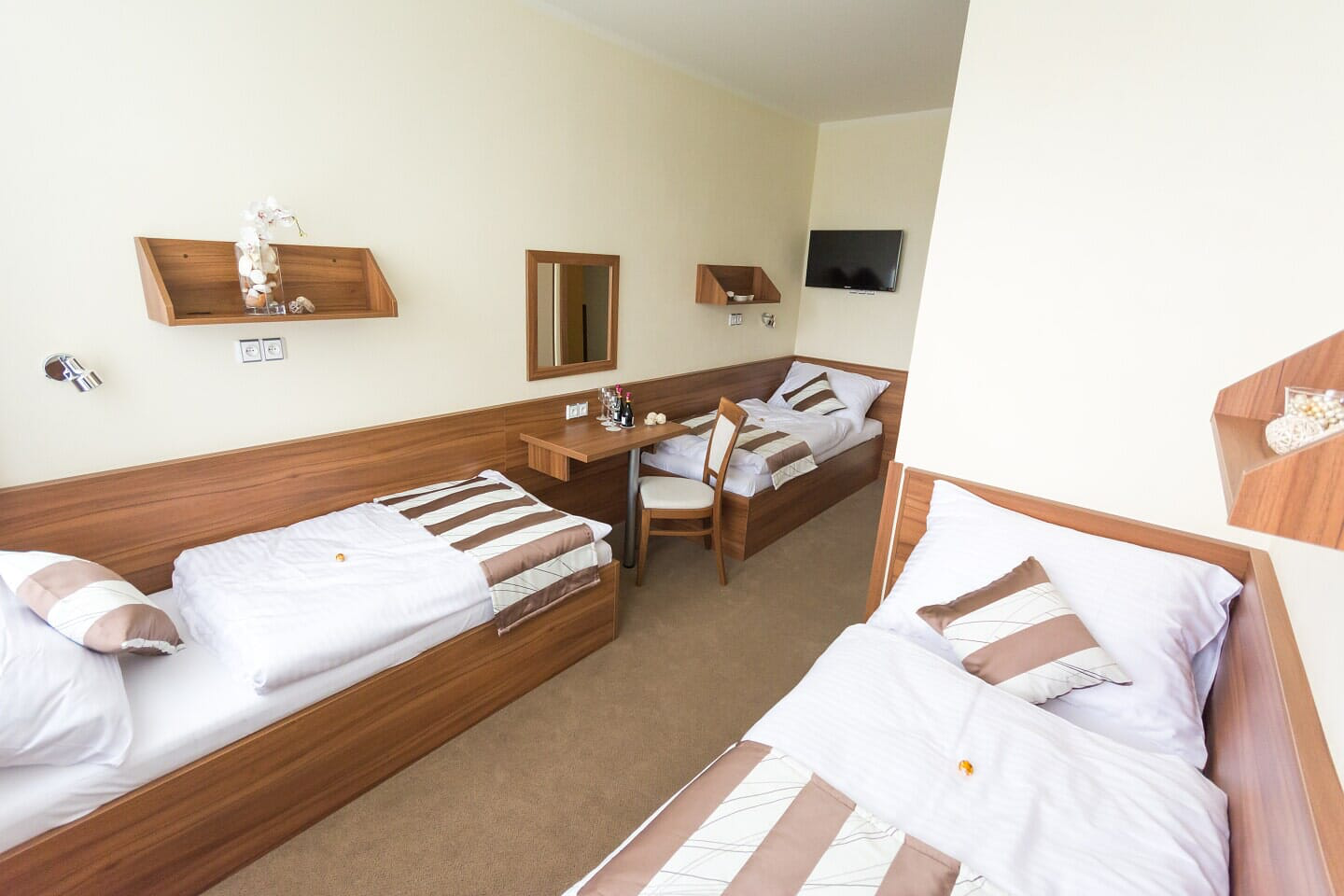 Ein Hotelzimmer mit drei Betten, einem Schreibtisch, einem Spiegel und einem kleinen Fernseher. Braun-weiße Einrichtung.
