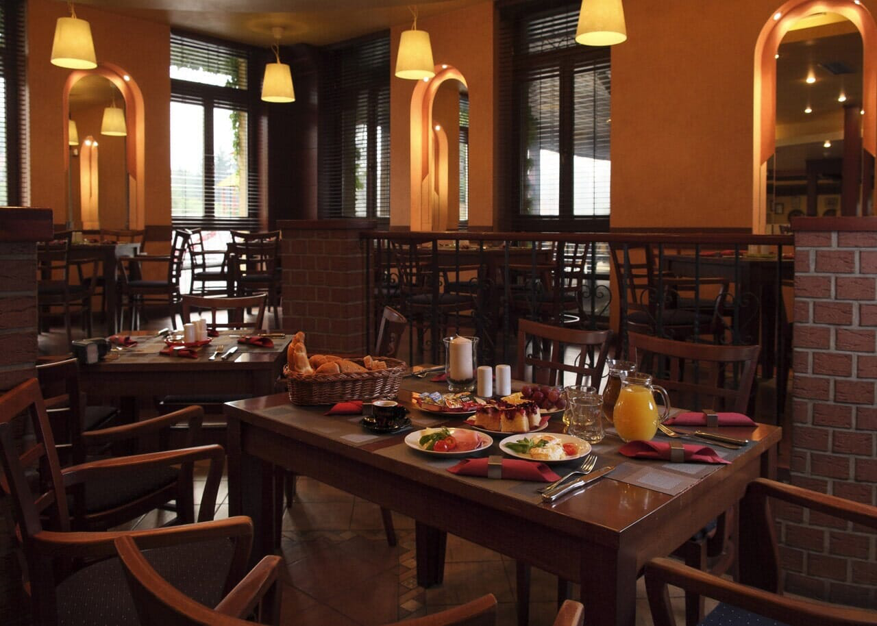 Restaurantinterieur mit zum Frühstück gedeckten Tischen. Braune Holzmöbel, orange Wände und natürliches Licht.