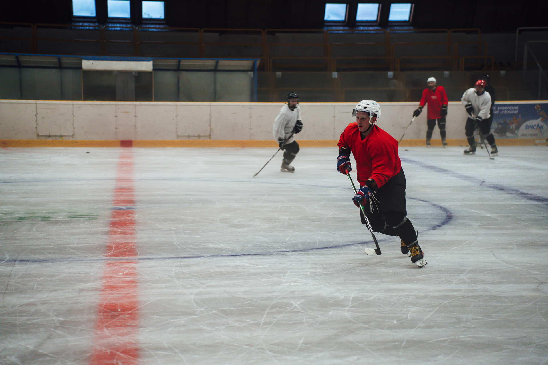 Ein Hockeyspieler im roten Trikot fährt mit dem Schläger in der Hand auf der Eisbahn Schlittschuh; im Hintergrund sind weitere Personen zu sehen.