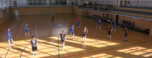 Handballspiel in einer Holzhalle. Die Spieler in blauen und schwarzen Trikots treten gegeneinander an.