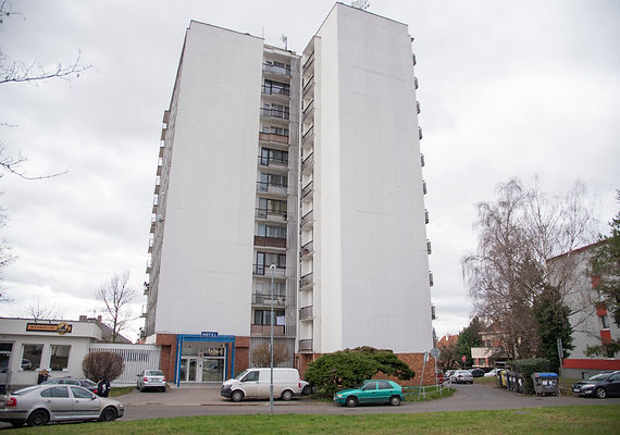 Weißes Hochhaus-Wohngebäude mit Balkonen, Eingang und davor geparkten Autos an einem bewölkten Tag.