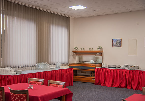 Buffetraum mit roten Tischdecken, Vitrine und Fenster mit Jalousien.