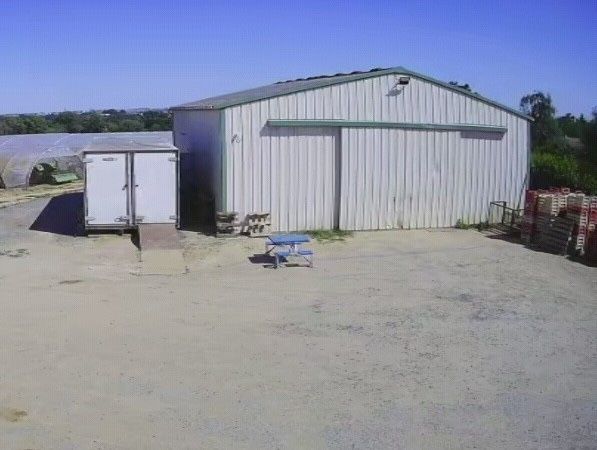 Petit hangar métallique blanc dans une cour poussiéreuse, avec une remorque blanche à gauche et des palettes empilées à droite.