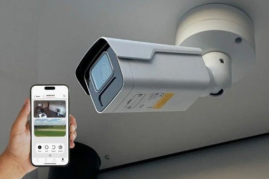 Main tenant un smartphone affichant le flux d'une caméra de sécurité à côté d'une caméra de vidéosurveillance blanche fixée au mur