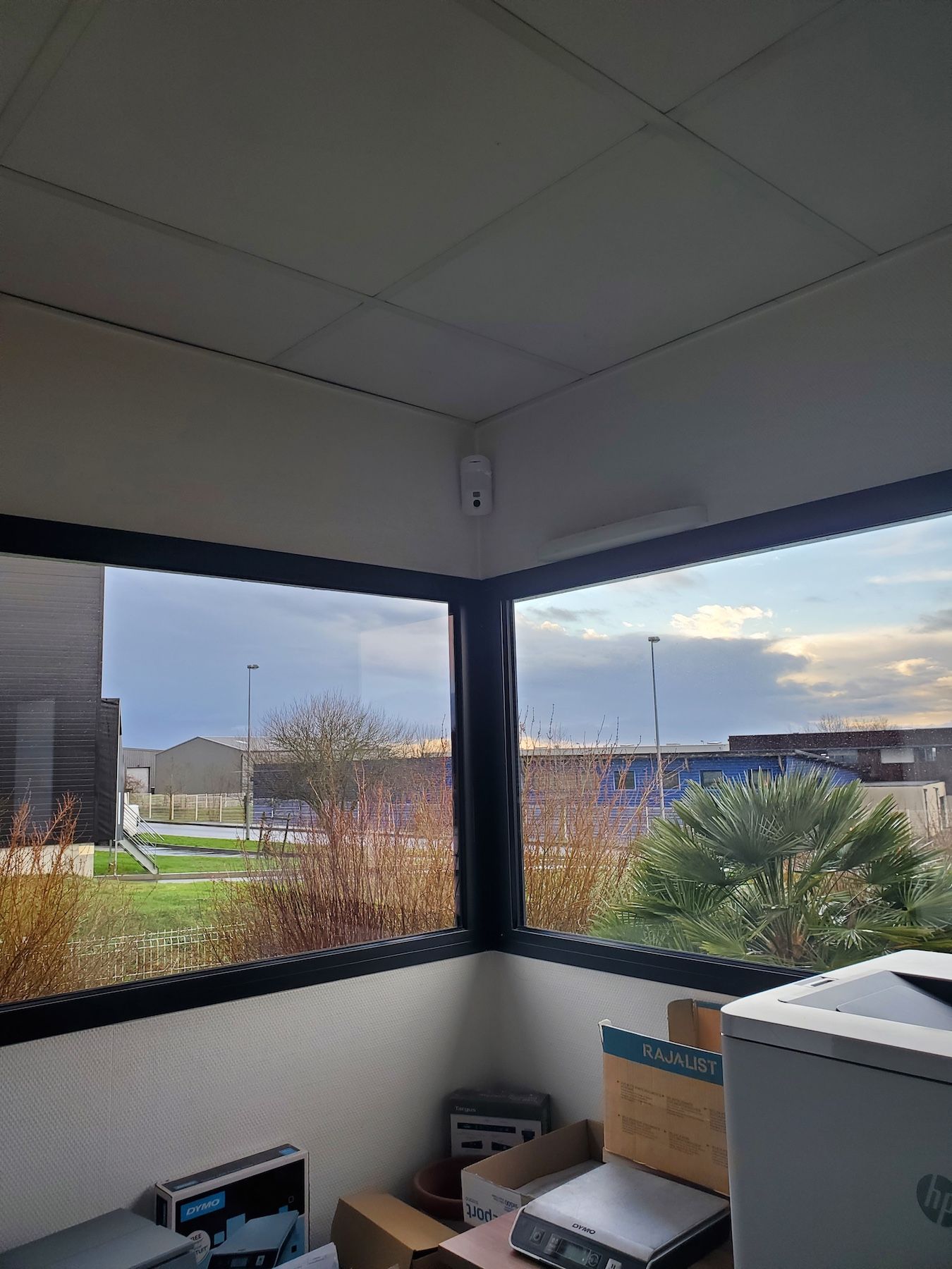 Bureau d'angle avec de grandes fenêtres donnant sur un parking et des bâtiments, des cartons empilés en dessous.