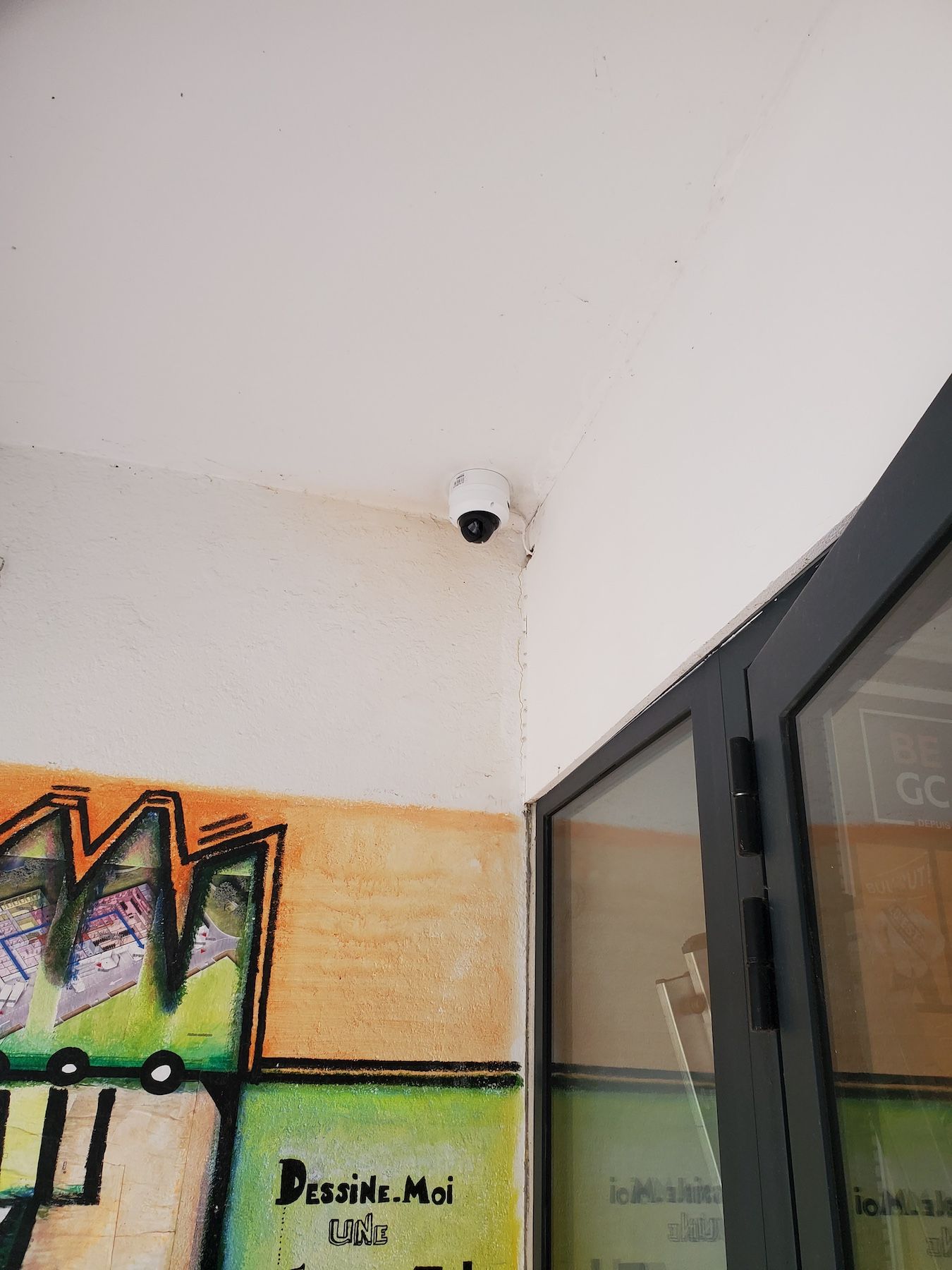 Coin d'une pièce avec des graffitis sur le mur et une petite caméra de sécurité au plafond près d'une porte vitrée ouverte.