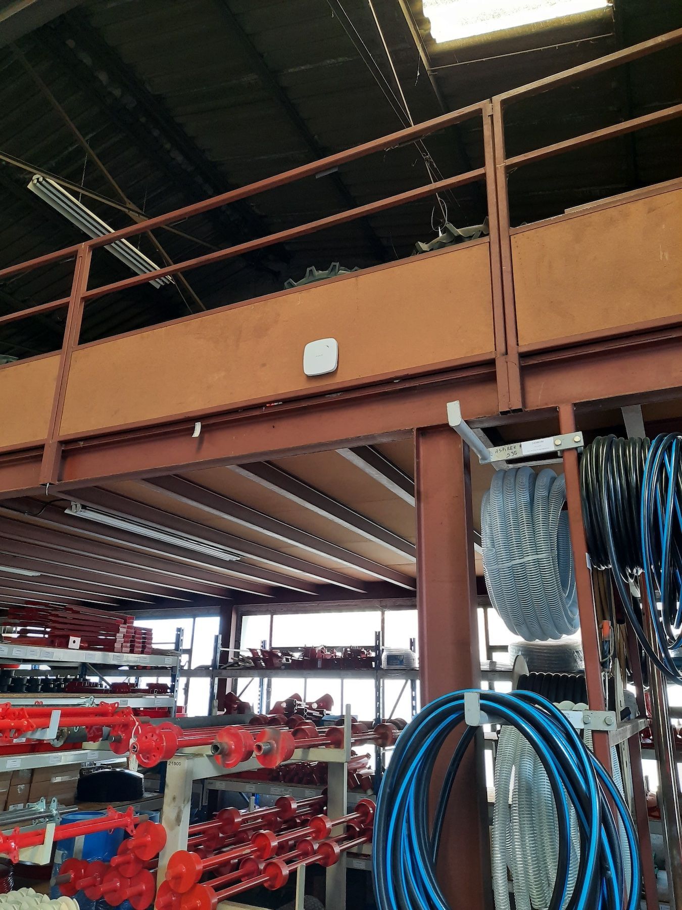 Zone de stockage industriel avec rayonnages métalliques, pièces rouges et tuyaux bleus enroulés sous une mezzanine.