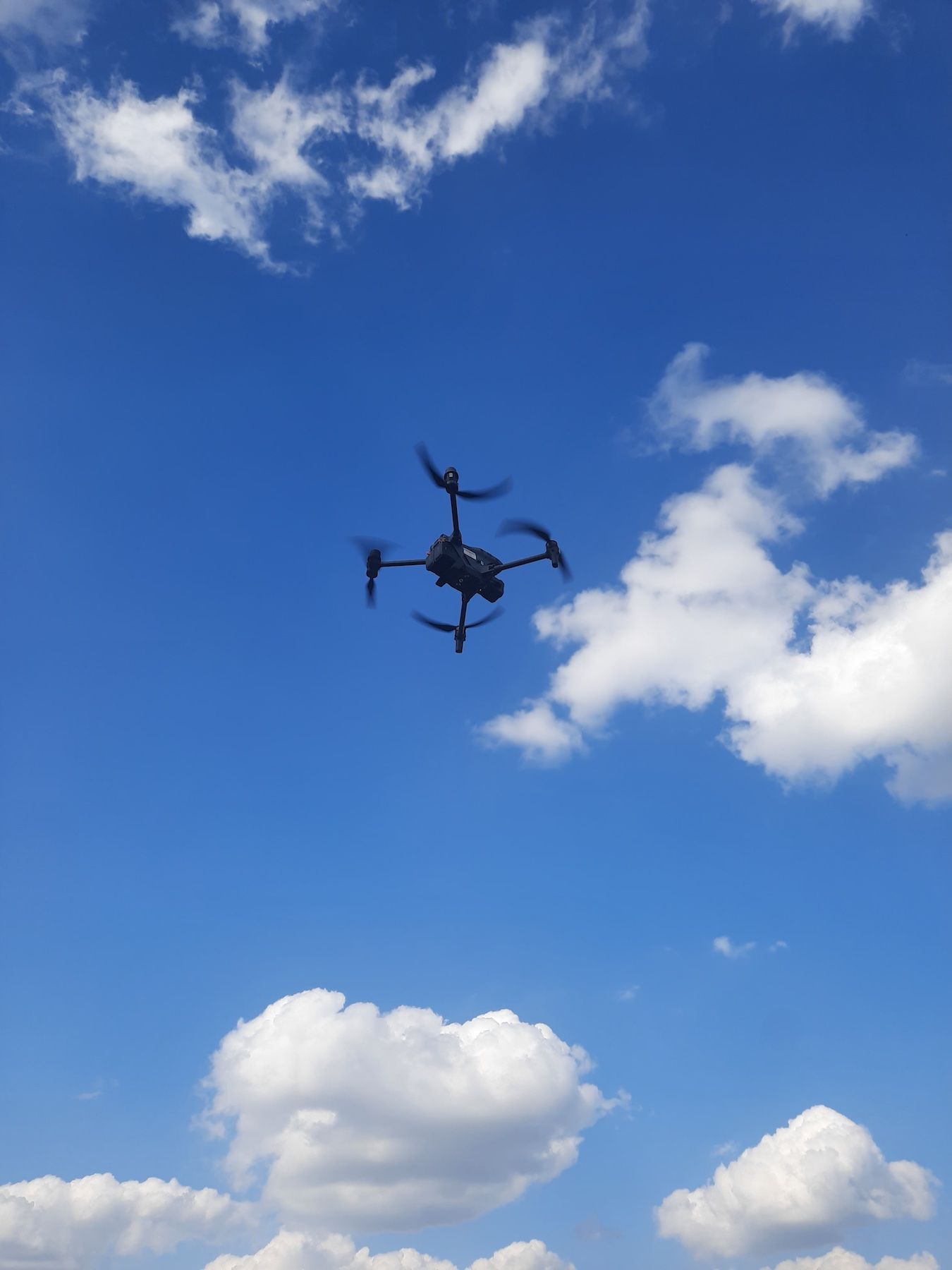 Un drone noir vole sur un ciel bleu clair parsemé de nuages ​​blancs.