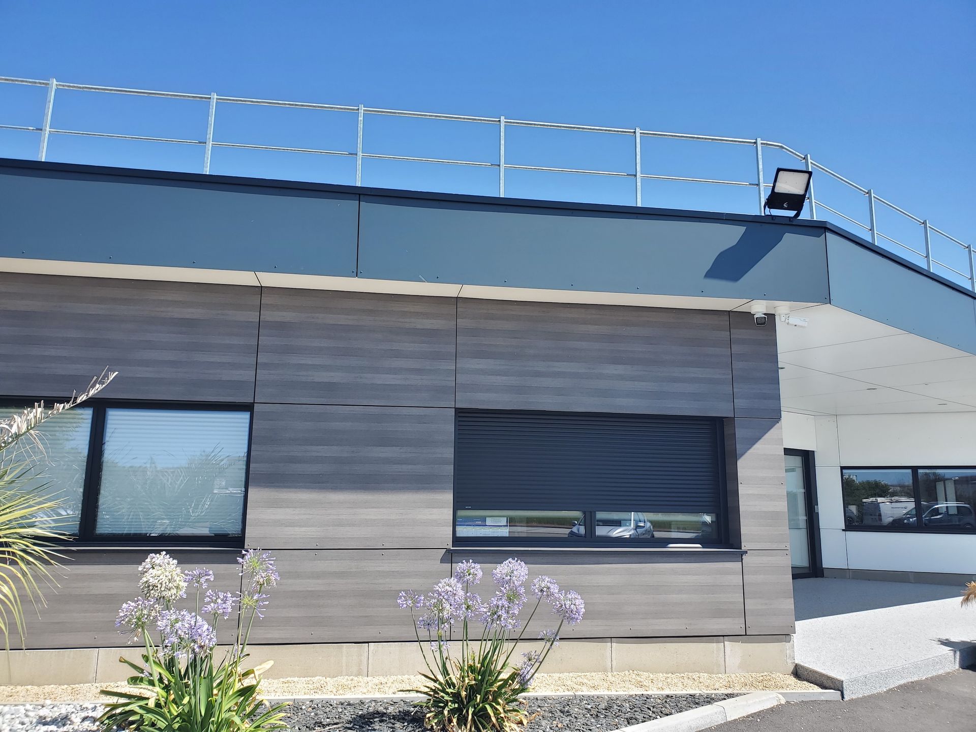 Extérieur d'un bâtiment moderne avec un bardage gris, des fenêtres sombres et une balustrade de toit bleue sous un ciel dégagé