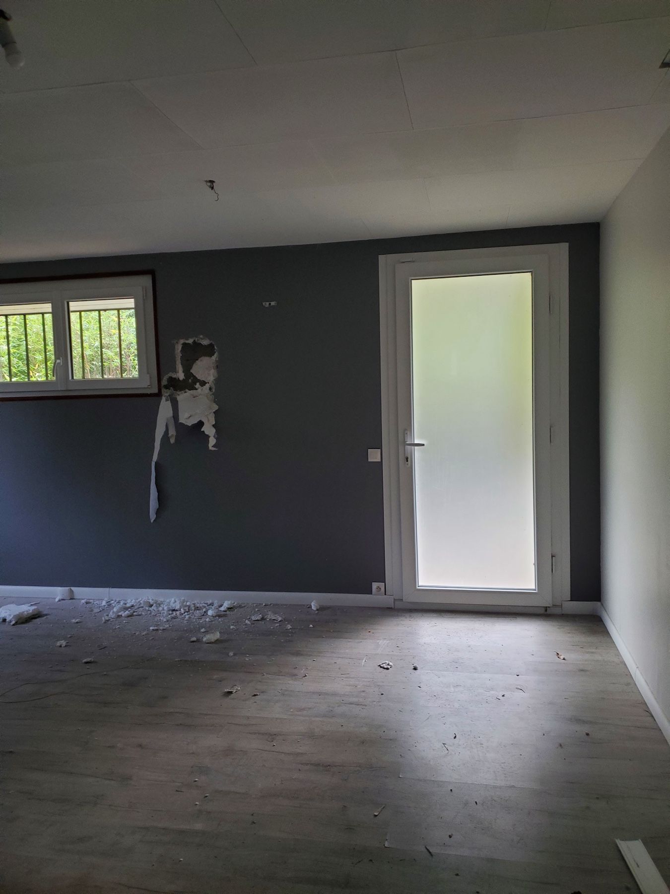 Pièce vide aux murs gris, sol nu, petite fenêtre et porte en verre dépoli.
