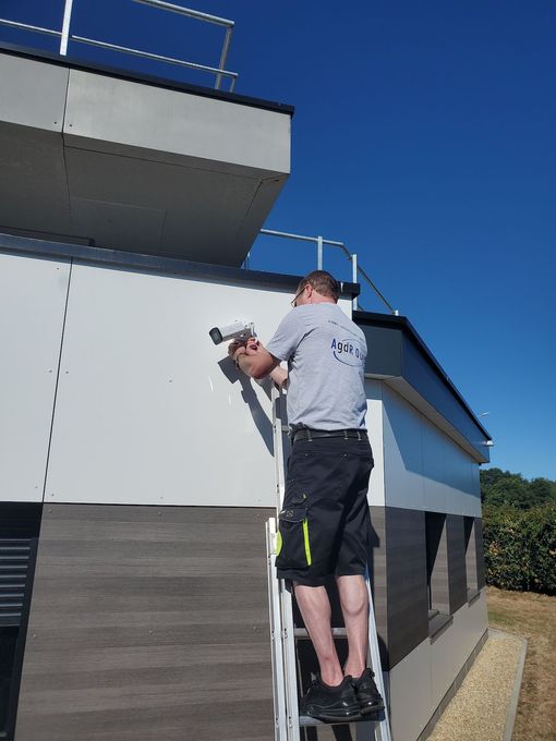 Un ouvrier utilise une perceuse sur la façade d'un bâtiment, debout sur une échelle sous un ciel bleu.
