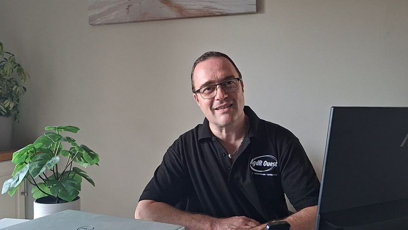 Un homme souriant, assis à un bureau avec un ordinateur portable, dans un bureau lumineux, avec une plante en pot à proximité.