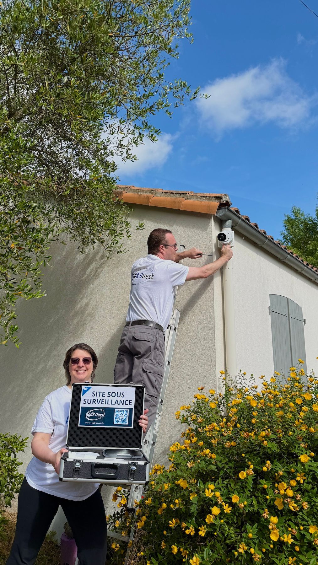 Deux personnes peignent l'extérieur d'une maison, l'une sur une échelle et l'autre tenant du matériel devant un buisson en fleurs.