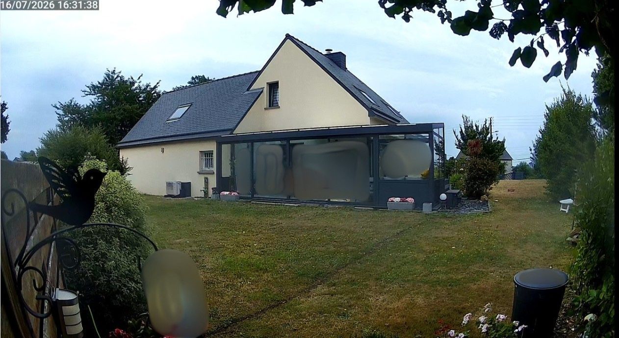 Jardin avec une maison couleur crème, un toit gris et une terrasse couverte sous un ciel couvert.