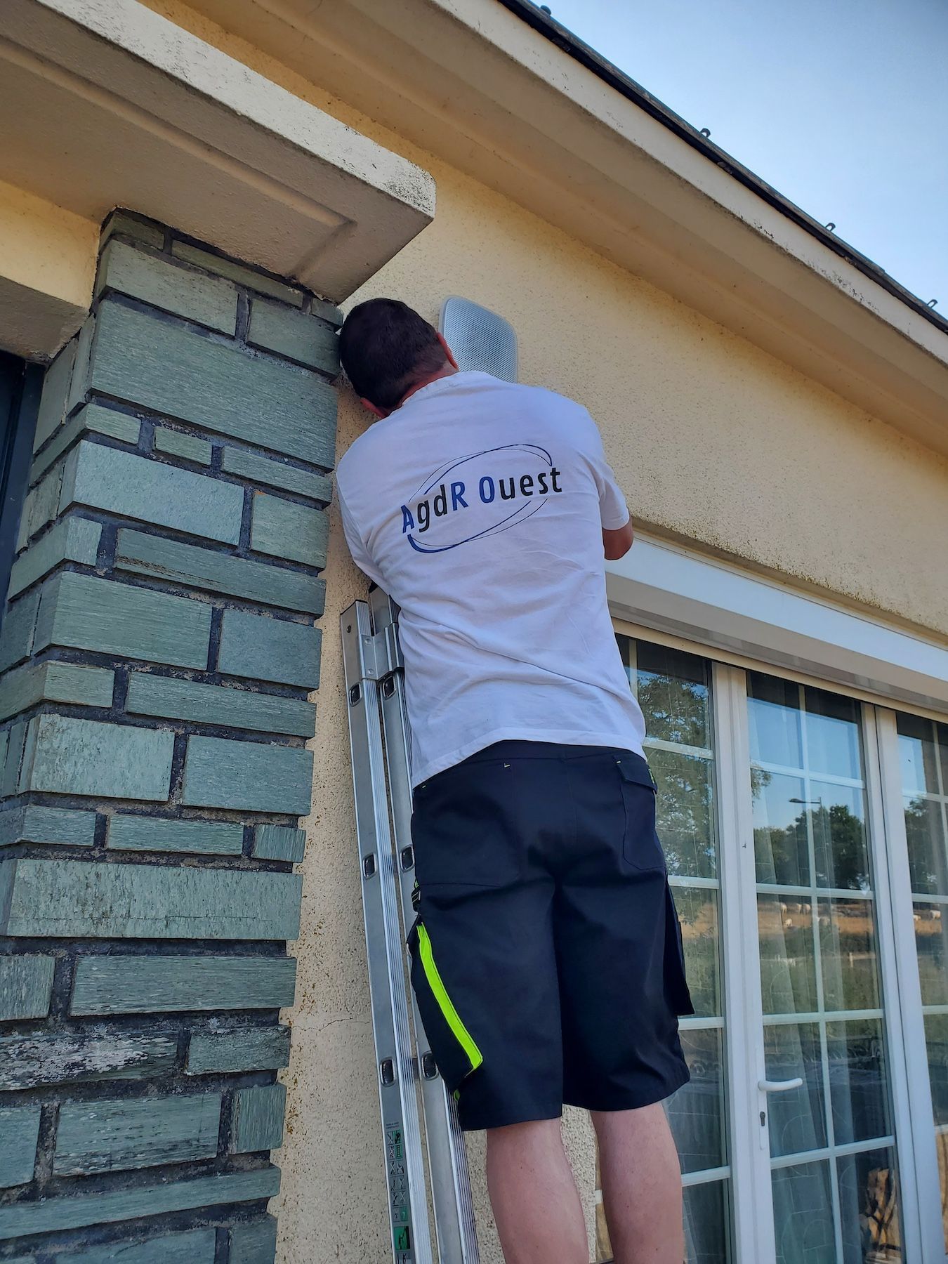 Une personne nettoie l'extérieur d'une maison à côté d'une colonne en briques et d'une fenêtre, à l'aide d'une échelle.