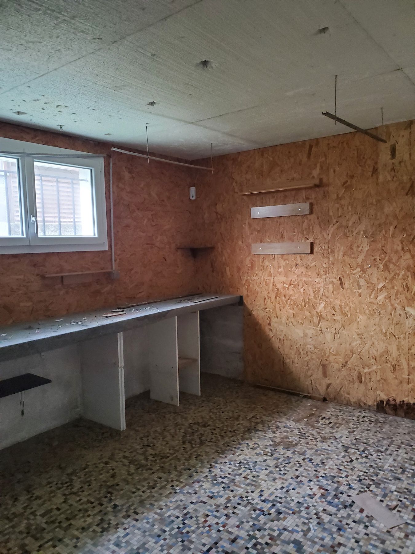 Pièce vide et inachevée avec des murs en panneaux OSB, une petite fenêtre, un comptoir intégré et un sol moucheté.