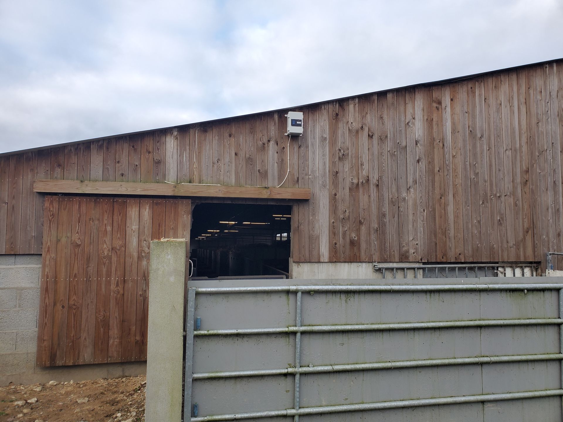 Grange en bois avec une porte ouverte derrière une clôture métallique grise sous un ciel couvert