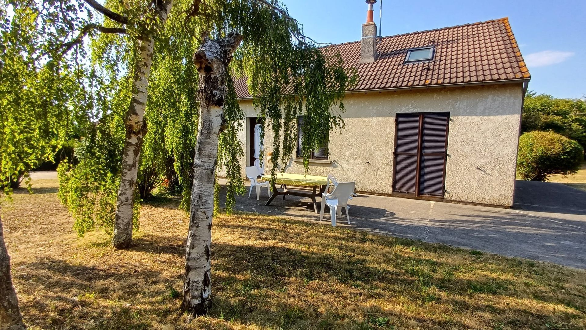Maisonnette en pierre au toit de tuiles rouges dans une cour ensoleillée, encadrée par un arbre feuillu au premier plan