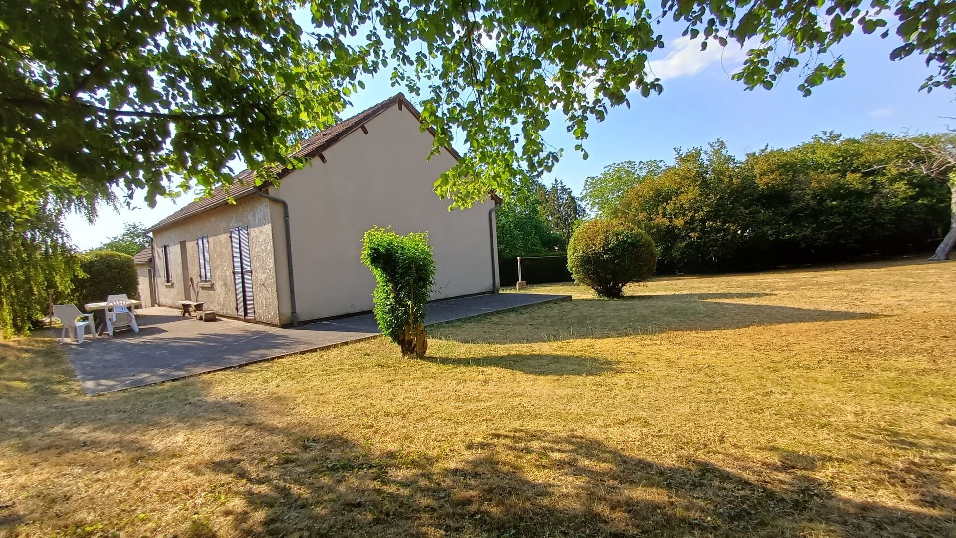 Maison de plain-pied avec un toit en pente, pelouse jaune sèche et arbres sous un ciel bleu