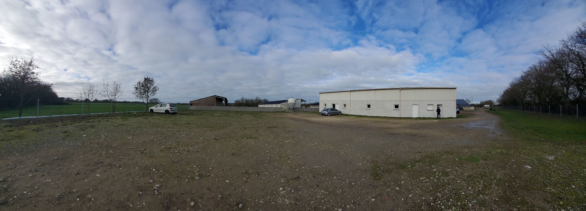Vaste terrain vague avec un petit bâtiment industriel blanc sous un ciel bleu nuageux.